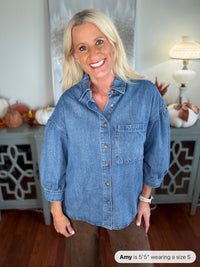 A woman wearing an oversized denim button-up shirt in medium blue with a chest pocket, styled with brown pants. The shirt features long sleeves and a classic collared design.