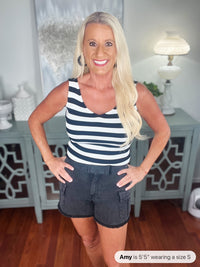 A woman wearing a navy and white striped sleeveless tank top paired with dark denim shorts with cargo pockets, standing indoors with hands on hips.