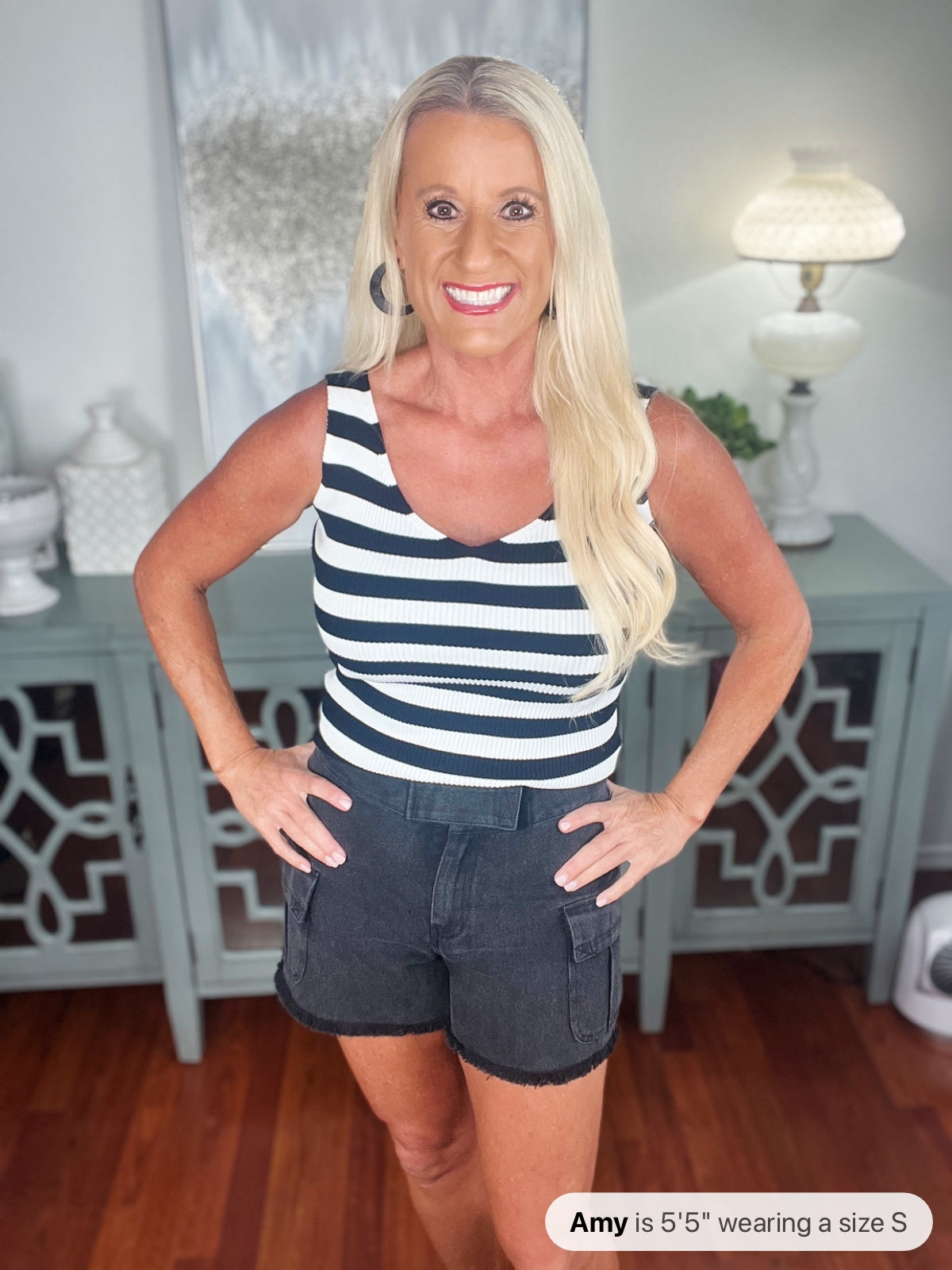 A woman wearing a navy and white striped sleeveless tank top paired with dark denim shorts with cargo pockets, standing indoors with hands on hips.