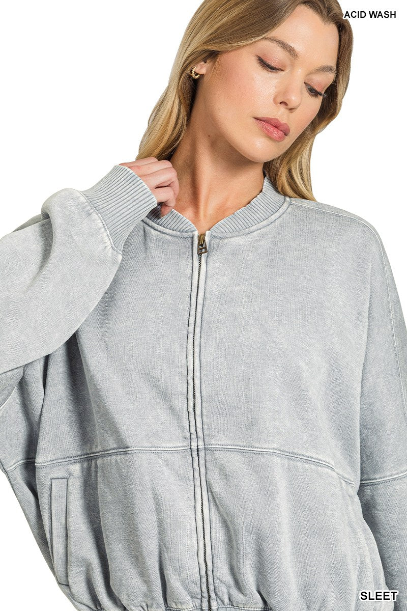 A woman wearing a light gray acid-washed zip-up sweatshirt with ribbed collar and cuffs, featuring a drawstring waist and front pockets.
