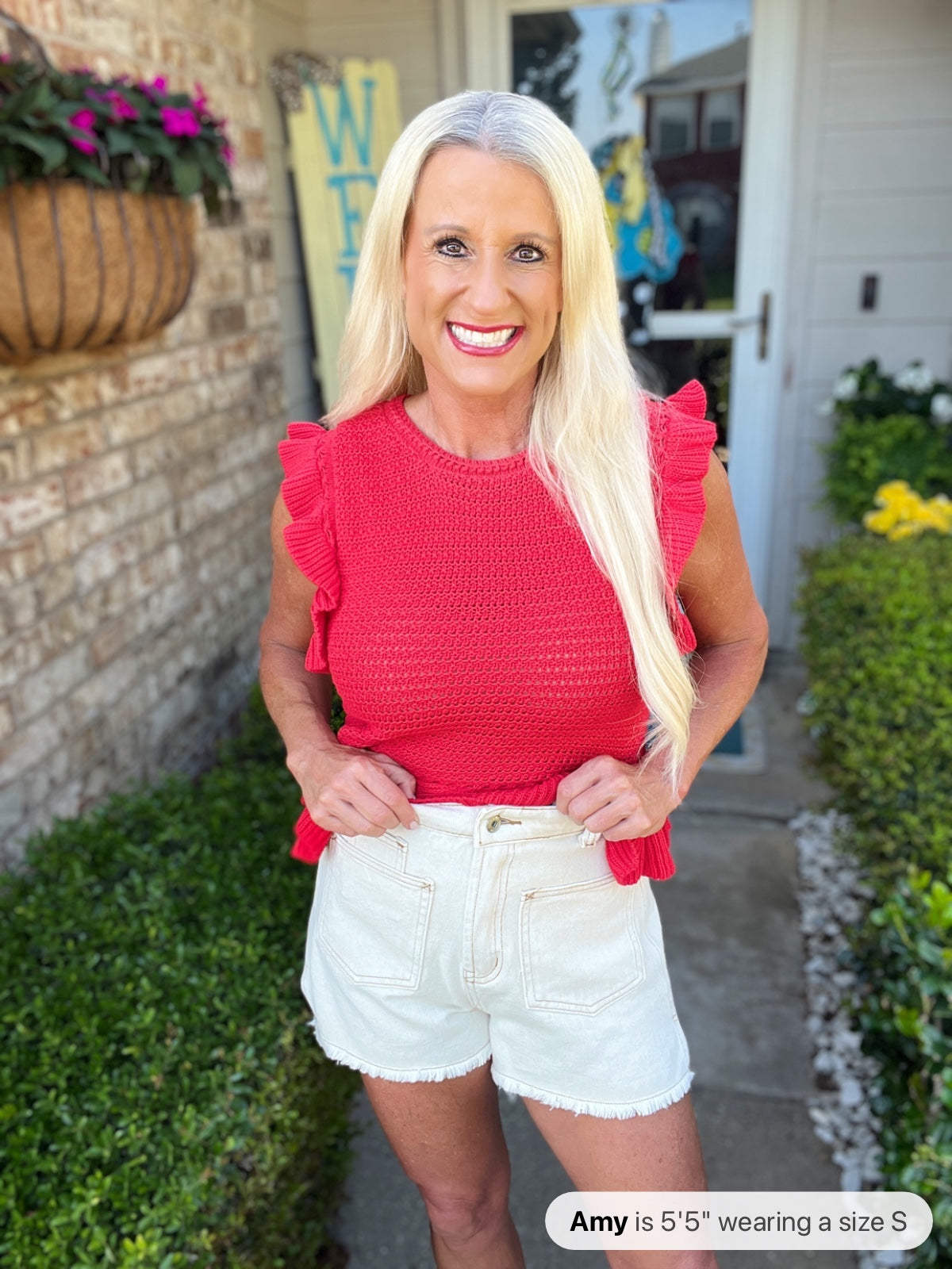 Woman wearing a bright coral pink sleeveless top with ruffled flutter cap sleeves and white denim shorts with a frayed hem, standing in front of a residential home entryway.