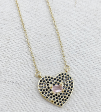 A gold-tone heart-shaped pendant necklace featuring black rhinestone pave detailing with a pale pink gemstone center, suspended from a delicate gold chain.