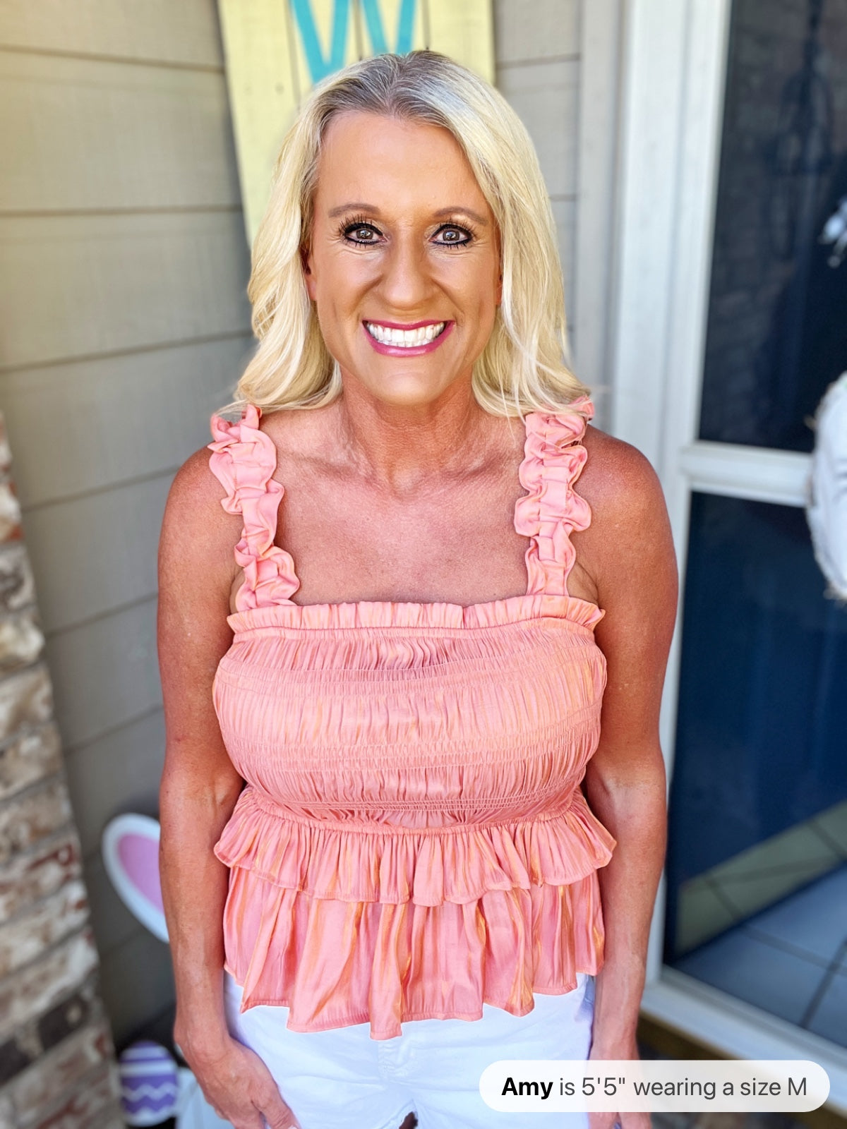 A woman wearing a coral pink sleeveless top with ruched texture, tiered ruffled details, and ruffled shoulder straps, paired with white pants.