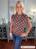 A woman wearing a brown sleeveless sweater vest with white polka dots, styled with blue jeans and layered pearl bracelets. The knit top features a classic crew neckline and relaxed fit.