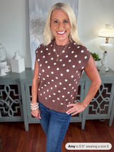 A woman wearing a brown sleeveless sweater vest with white polka dots, styled with blue jeans and layered pearl bracelets. The knit top features a classic crew neckline and relaxed fit.
