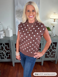 A woman wearing a brown sleeveless sweater vest with white polka dots, styled with blue jeans and layered pearl bracelets. The knit top features a classic crew neckline and relaxed fit.