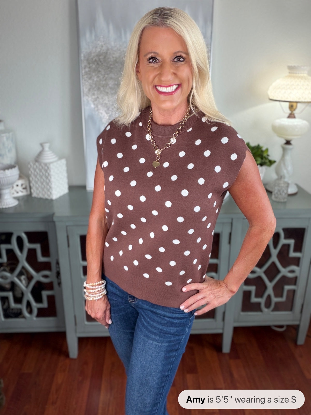 A woman wearing a brown sleeveless sweater vest with white polka dots, styled with blue jeans and layered pearl bracelets. The knit top features a classic crew neckline and relaxed fit.