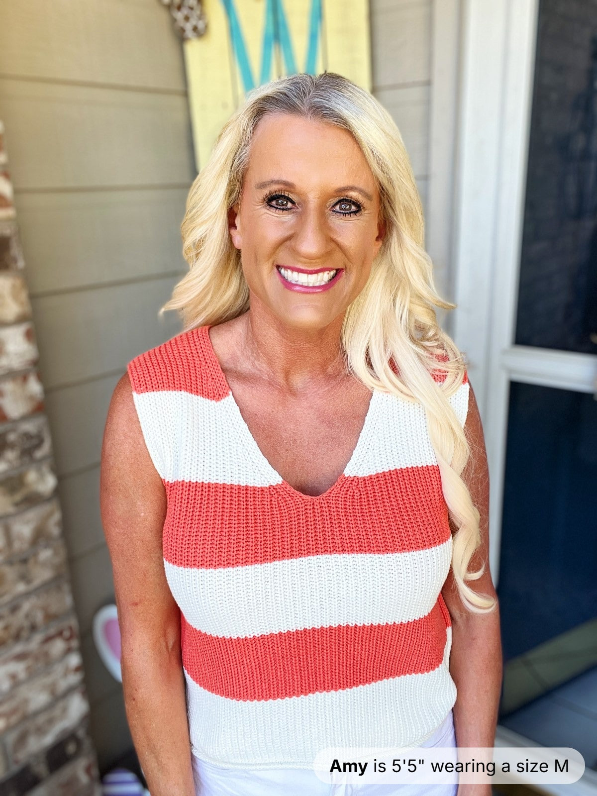 A woman wearing a sleeveless striped knit tank top in coral red and white horizontal stripes with a V-neckline, paired with white pants.