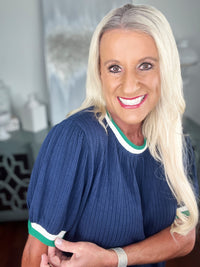A woman wearing a navy blue ribbed knit sweater with white and green striped trim at the neckline and short sleeves, paired with a silver bracelet.