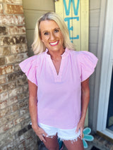 A woman wearing a pink and white striped short-sleeve blouse with ruffled flutter sleeves and a V-neckline, paired with white distressed denim shorts.