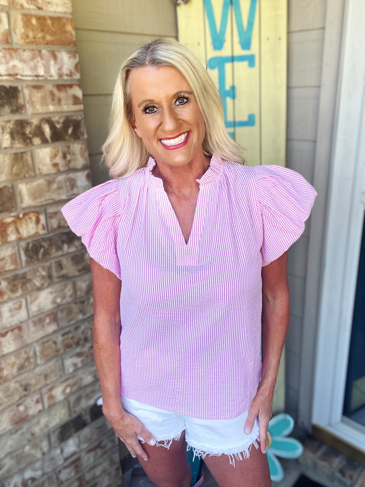 A woman wearing a pink and white striped short-sleeve blouse with ruffled flutter sleeves and a V-neckline, paired with white distressed denim shorts.