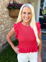 A woman wearing a bright coral red sleeveless sweater with ruffled flutter sleeves and a pleated hem, paired with white shorts. The textured knit top features a crew neckline and feminine ruffle details on the shoulders.