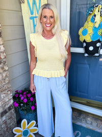 A woman wearing a pale yellow sleeveless knit top with ruffled hem paired with light blue wide-leg pants, styled for a casual spring look.