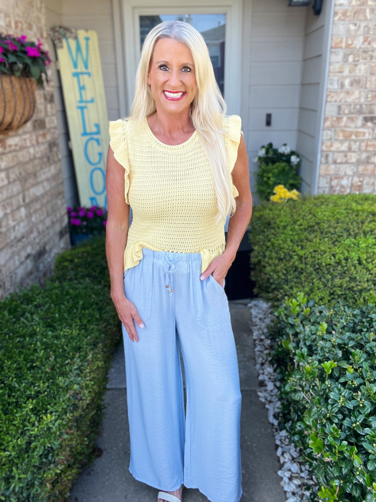 A woman wears a yellow sleeveless knit top with ruffled cap sleeves paired with light blue wide-leg pants. She is standing in front of a brick home entrance with a "Welcome" sign and flowering planters.