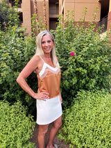 A woman wearing a two-tone sleeveless dress with a tan/caramel colored top and white bottom, featuring a white trim detail at the neckline, poses in a garden setting surrounded by green foliage and pink flowers.