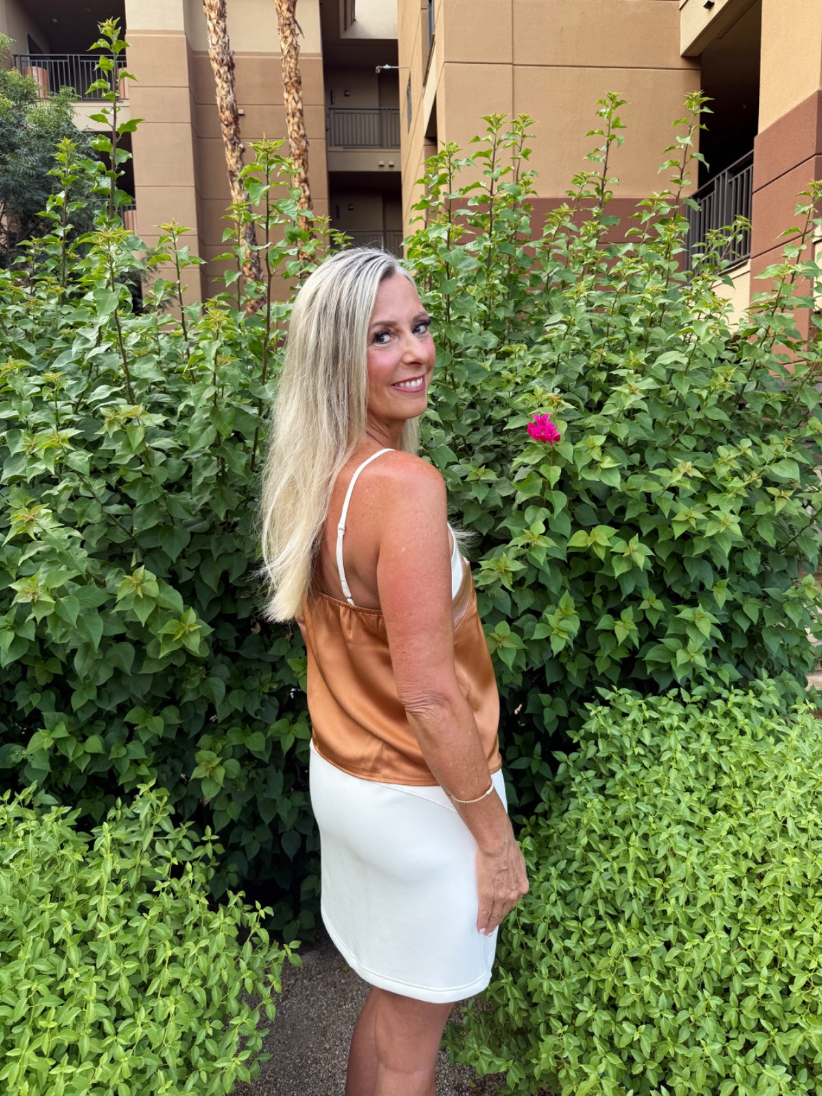 Woman wearing a bronze/tan sleeveless tank top with white trim and a white skirt, standing among lush green plants and flowers in an outdoor garden setting.