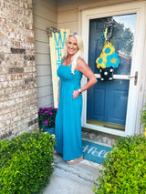 A woman wearing a bright turquoise sleeveless maxi dress with a halter neckline stands on a front porch. The dress features a relaxed, flowing silhouette and is styled casually for summer wear.