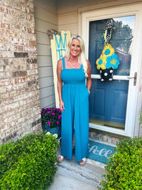 A woman wearing a bright blue sleeveless empire-waist maxi dress stands on a front porch decorated with spring flowers and seasonal door decorations. The dress features a gathered bodice and flowing skirt with a comfortable, casual silhouette.