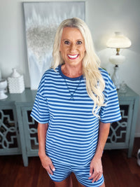 A woman wearing a blue and white striped short-sleeve t-shirt paired with matching striped shorts, styled with a layered necklace in a bright, modern interior setting.