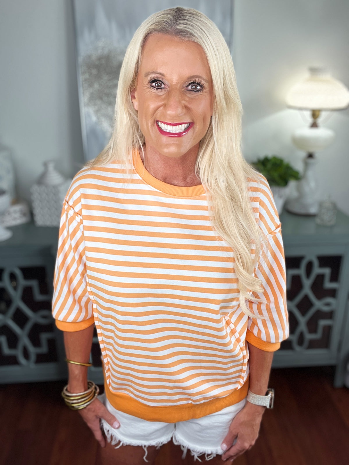 Woman wearing an orange and white striped three-quarter sleeve crew neck sweater paired with white distressed denim shorts, accessorized with gold bangles.