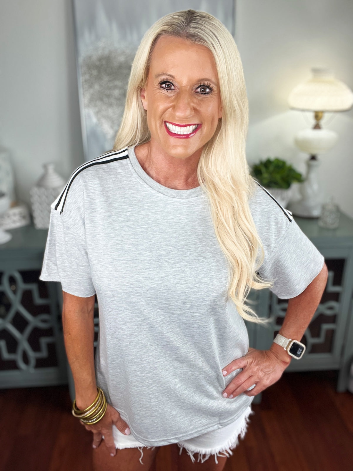 Woman wearing a light gray short-sleeve t-shirt with black stripes on the shoulders, paired with white distressed shorts. She is smiling at the camera in an indoor setting.