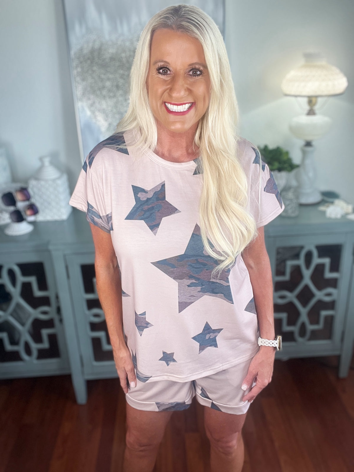 Woman wearing a light pink short-sleeve t-shirt with navy blue star print pattern, paired with matching pink shorts. The casual outfit is styled with a bracelet and displayed against a light home interior background.
