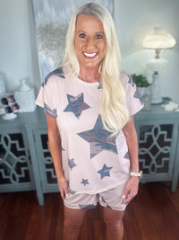 Woman wearing a light pink short-sleeve t-shirt with navy blue star print pattern, paired with matching pink shorts. The casual outfit is styled with a bracelet and displayed against a light home interior background.