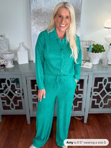 A woman wearing a teal ribbed button-up jumpsuit with long sleeves and a drawstring waist, styled with white sneakers in a home interior setting.