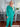 A woman wearing a teal ribbed button-up jumpsuit with long sleeves and a drawstring waist, styled with white sneakers in a home interior setting.