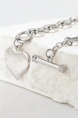 A silver toggle bracelet featuring a chunky chain link design with a polished heart charm pendant and cylindrical toggle clasp, displayed on a white textured stone surface.