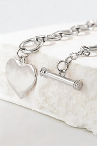 A silver toggle bracelet featuring a chunky chain link design with a polished heart charm pendant and cylindrical toggle clasp, displayed on a white textured stone surface.