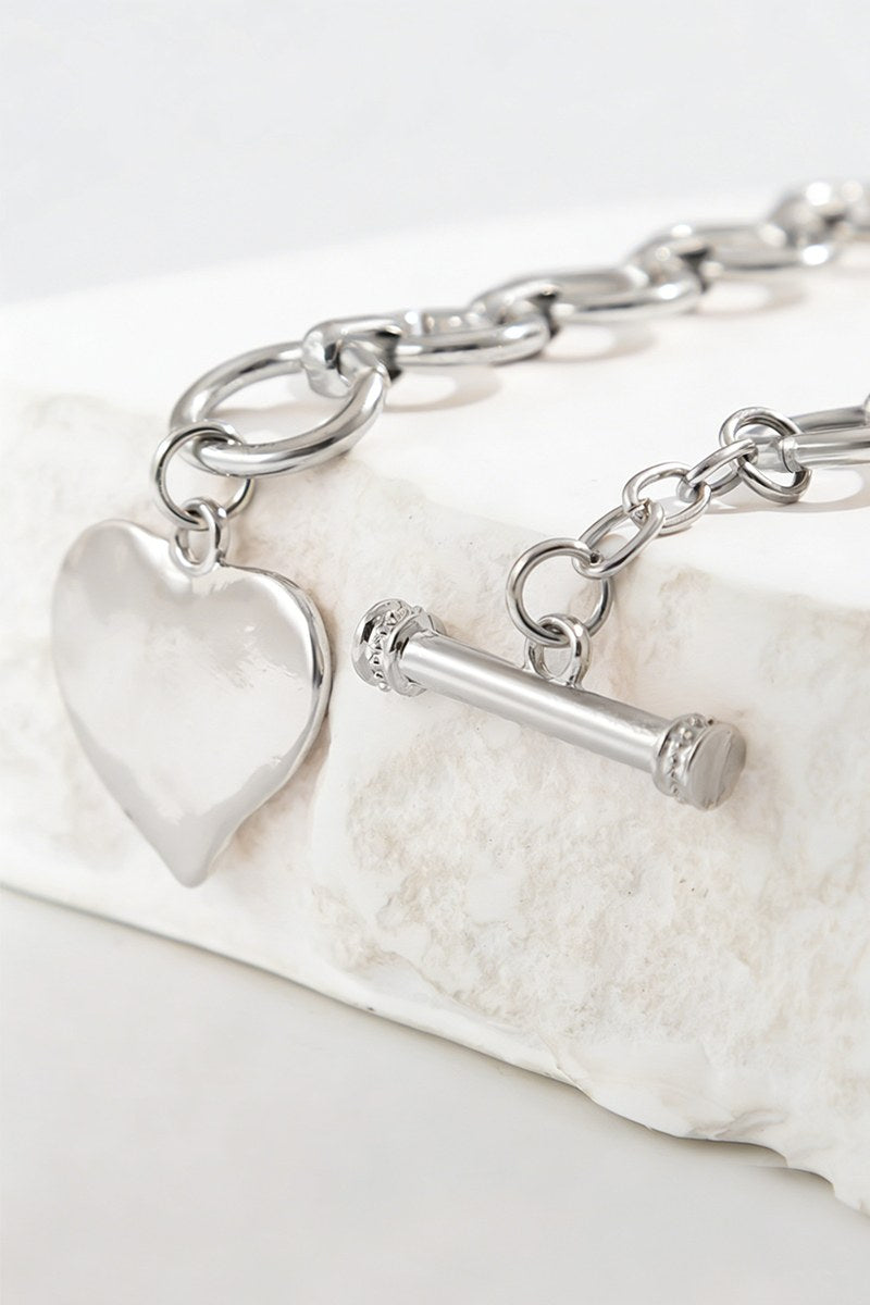 A silver toggle bracelet featuring a chunky chain link design with a polished heart charm pendant and cylindrical toggle clasp, displayed on a white textured stone surface.