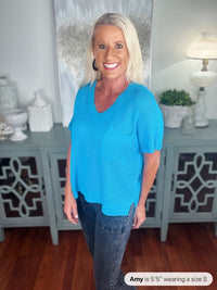A woman wearing a bright turquoise ribbed short-sleeve v-neck sweater paired with dark wash jeans, standing in front of a gray cabinet in a bright home interior.