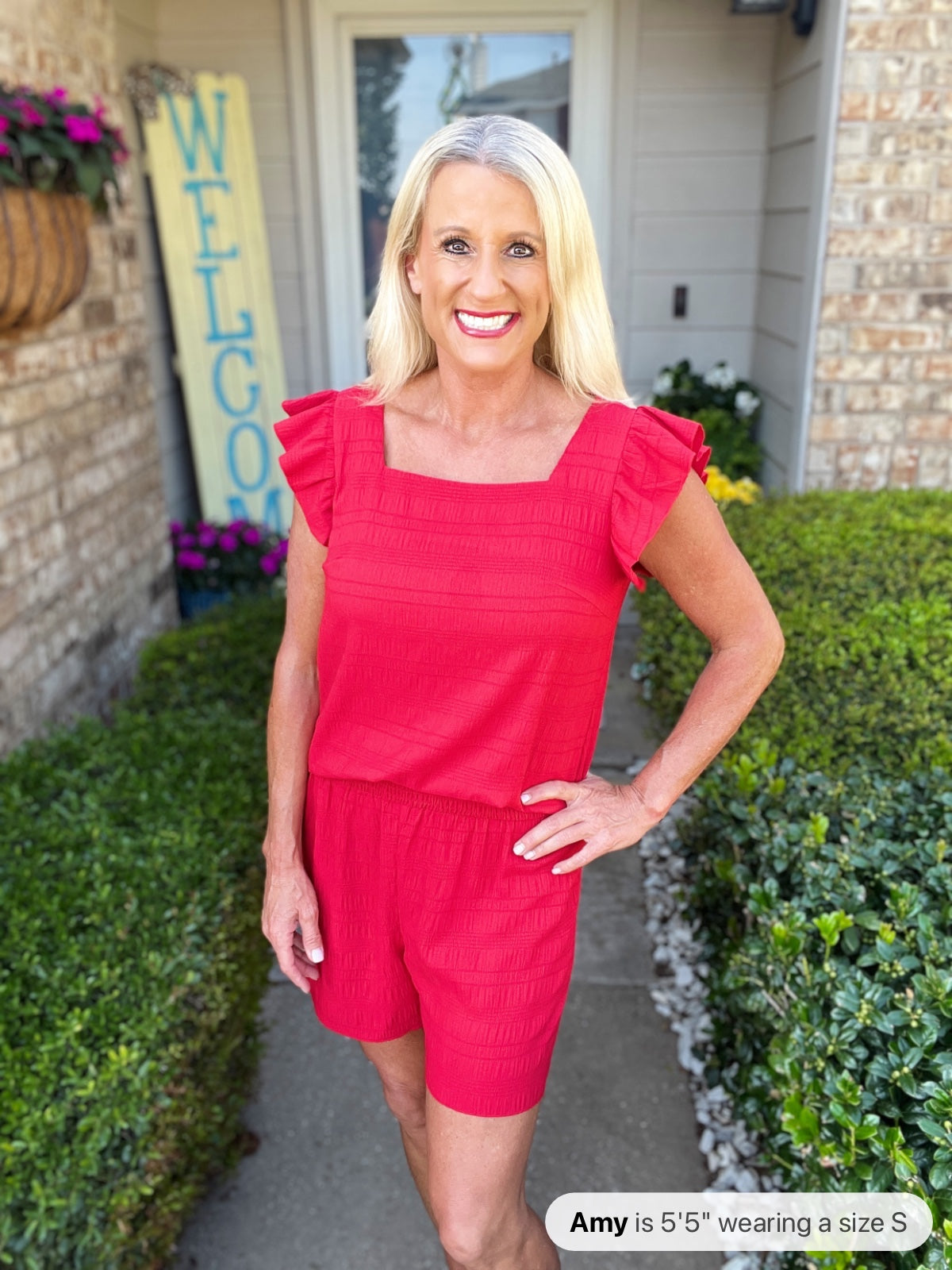 Woman wearing a bright red textured romper with ruffled flutter sleeves and shorts-length legs, standing in front of a brick home entrance with a "Welcome" sign.
