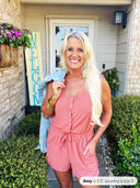 A woman wearing a pink sleeveless romper with button-front closure and drawstring waist, styled with gold bangles and long blonde hair, poses in front of a home entrance.