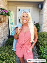 A woman wearing a pink sleeveless romper with button-front closure and drawstring waist, styled with gold bangles and long blonde hair, poses in front of a home entrance.