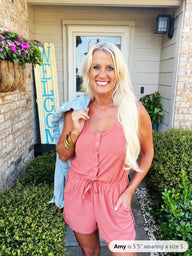 A woman wearing a pink sleeveless romper with button-front closure and drawstring waist, styled with gold bangles and long blonde hair, poses in front of a home entrance.