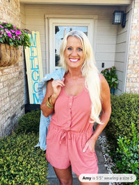 A woman wearing a pink sleeveless romper with button-front closure and drawstring waist, styled with gold bangles and long blonde hair, poses in front of a home entrance.