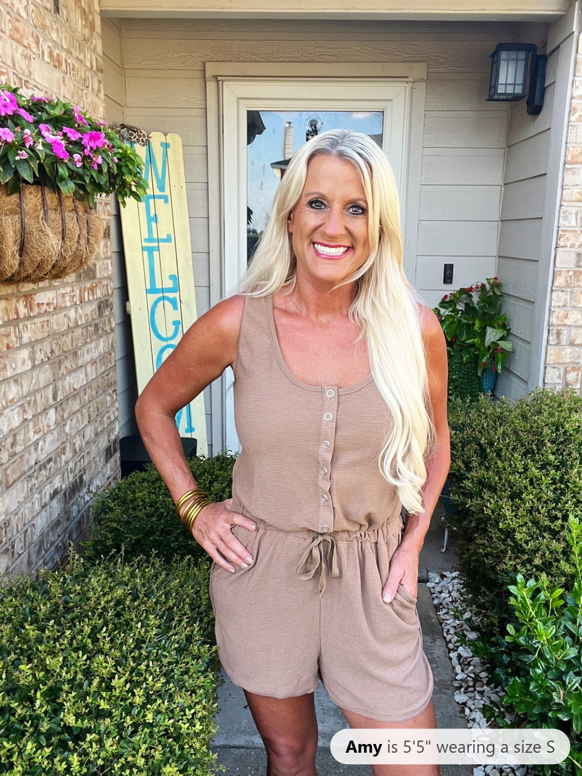 Woman wearing a beige sleeveless romper with button front and drawstring waist, paired with gold bangle bracelets, photographed on a residential porch.