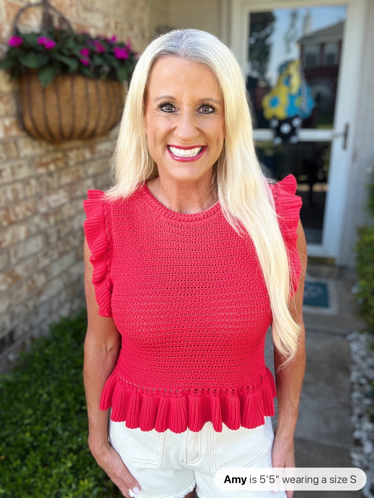 A woman wearing a bright red sleeveless top with ruched shoulders and a pleated ruffle hem, paired with white shorts. The textured knit top features a crew neckline and fitted silhouette.