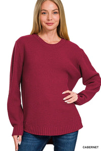 A woman wearing a cabernet-colored knit sweater with a textured pattern, long sleeves with ribbed cuffs, and a ribbed hem, paired with blue jeans.