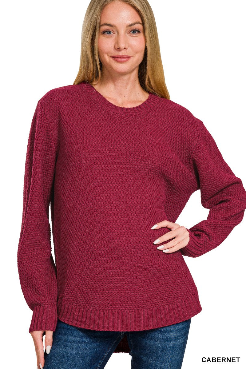 A woman wearing a cabernet-colored knit sweater with a textured pattern, long sleeves with ribbed cuffs, and a ribbed hem, paired with blue jeans.