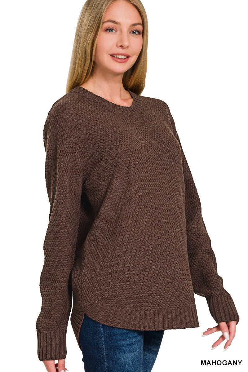 A woman wearing a mahogany brown textured knit sweater with long sleeves and ribbed cuffs, styled over dark blue jeans against a white background.