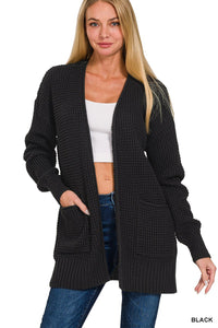 A woman wearing a black waffle-knit cardigan with front pockets over a white ribbed crop top and dark blue jeans. The cardigan features a relaxed fit with long sleeves and ribbed cuffs and hem.