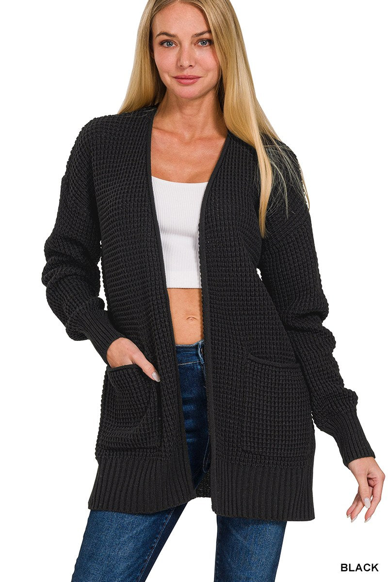 A woman wearing a black waffle-knit cardigan with front pockets over a white ribbed crop top and dark blue jeans. The cardigan features a relaxed fit with long sleeves and ribbed cuffs and hem.