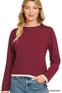 A woman wearing a dark burgundy long-sleeve crop sweater with a crew neckline and contrasting cream-colored trim, paired with blue jeans.