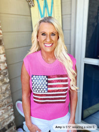 A woman wearing a bright pink sleeveless knit tank top featuring a sequined American flag design in red, white, and blue across the chest, paired with white pants.
