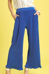 A woman wearing bright blue pleated wide-leg pants with a scalloped hem, paired with a cream-colored top and white platform sandals, standing against a yellow background.