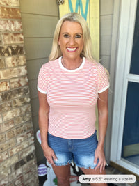Woman wearing a pink and white striped short-sleeve crew neck t-shirt paired with distressed denim shorts, standing in a home entryway.