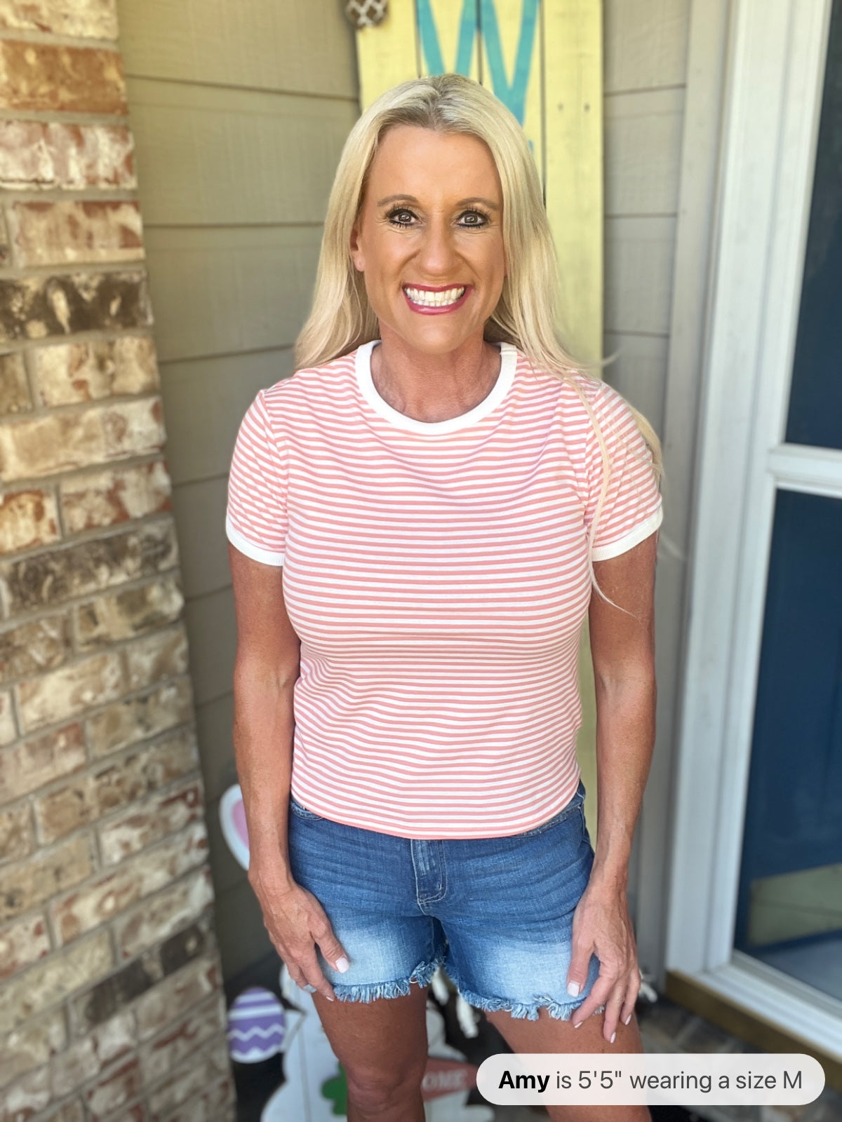Woman wearing a pink and white striped short-sleeve crew neck t-shirt paired with distressed denim shorts, standing in a home entryway.